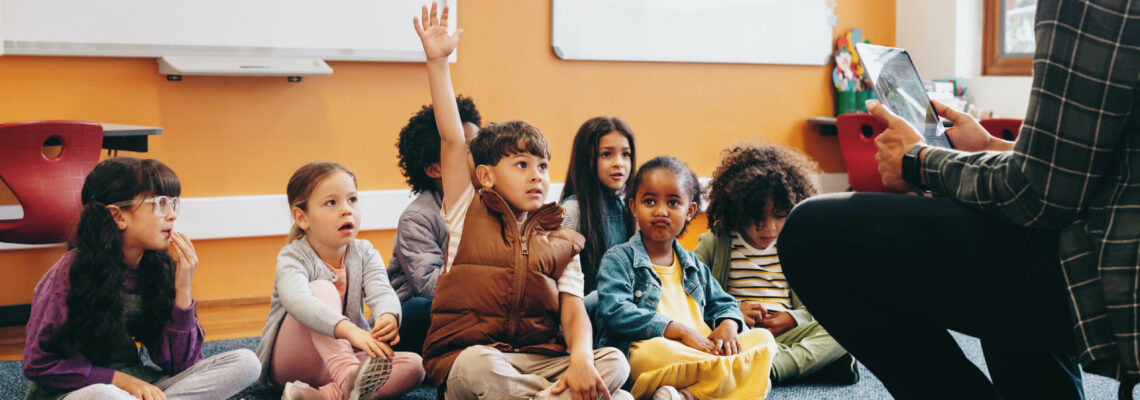 Boy raises his hand to answer a question in a classroom; he is sitting on the floor with other kids and the teacher is sitting in front of the class. Early child development in an elementary school.